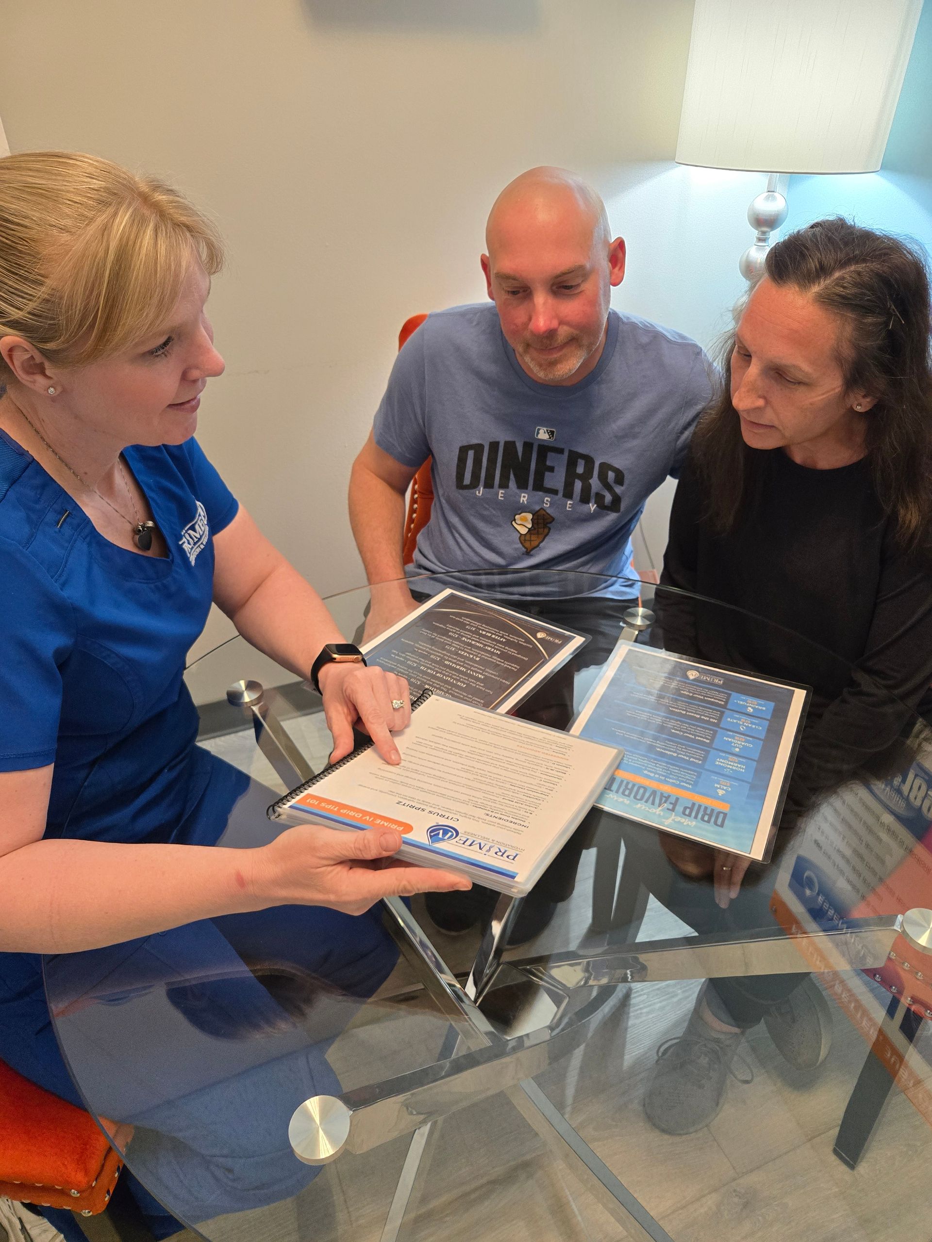 A Prime IV nurse in blue scrubs reviews a drip menu and wellness guide with two seated guests at a glass table